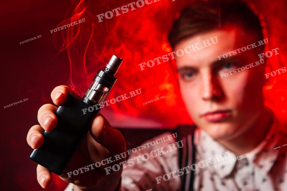 На фотографии изображен молодой человек, держащий вейп-устройство на фоне густого красного дыма. Снимок подчеркивает современную культуру вейпинга и стильный образ пользователя.